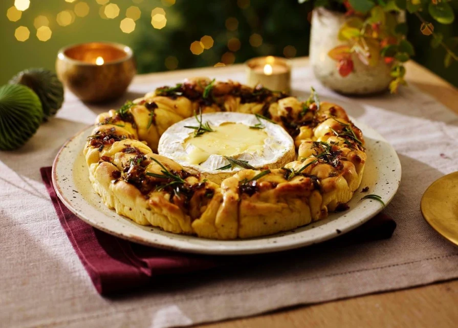 Overhead shot of brie in a bread wreath with herbs, served on a plate atop a red napkin, with candles and lights in background.