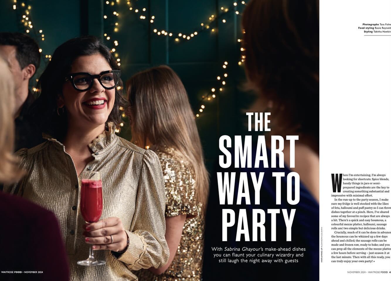 Smiling woman in a gold blouse holds a cocktail glass at a party with other guests, illuminated by string lights in the background.