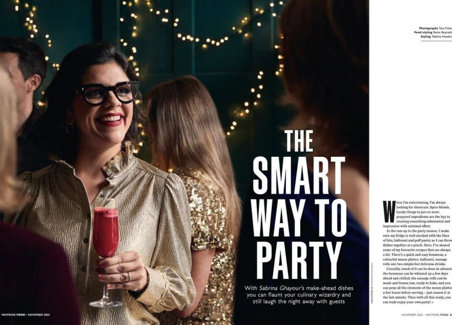 Smiling woman in a gold blouse holds a cocktail glass at a party with other guests, illuminated by string lights in the background.