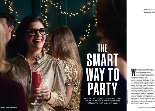 Smiling woman in a gold blouse holds a cocktail glass at a party with other guests, illuminated by string lights in the background.