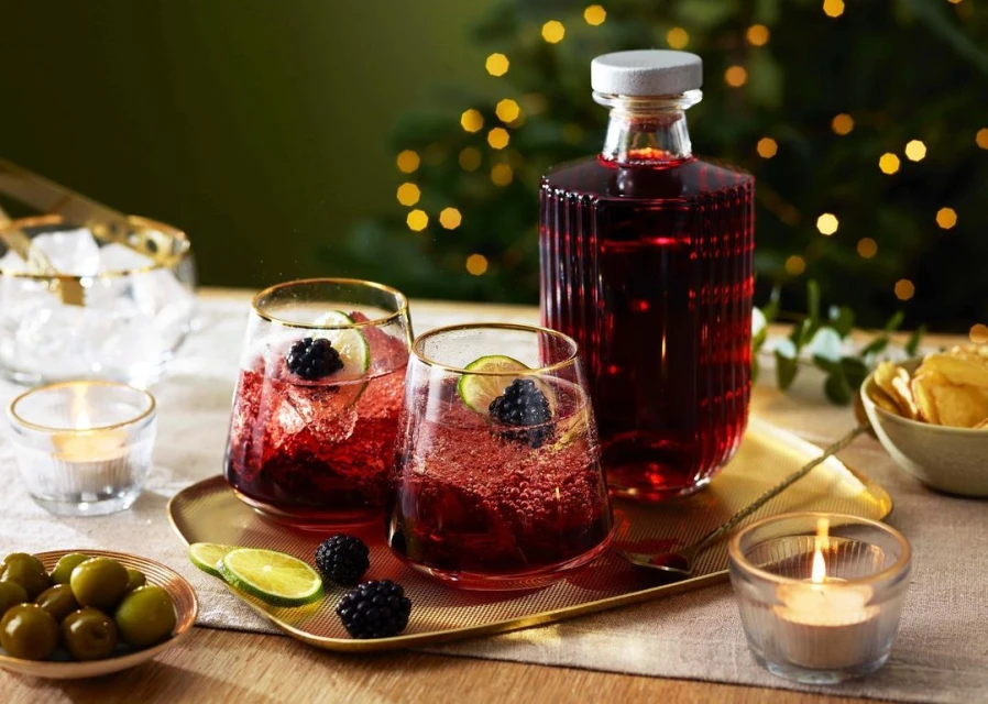 Two glasses filled with a red drink, lime, and blackberries on a table with a bottle, small dish of olives, and twinkling lights.