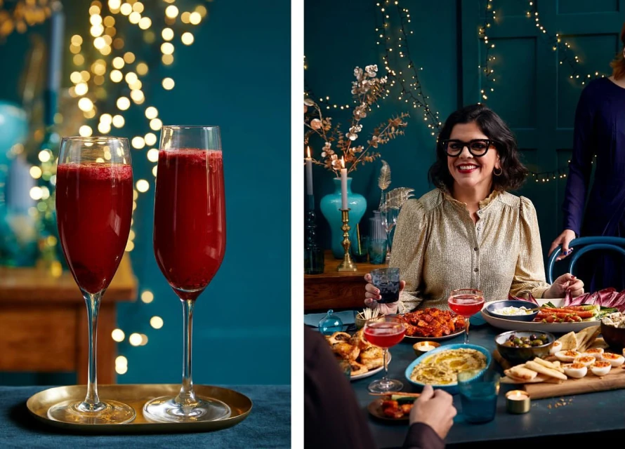 Two fluted glasses of red beverage are placed on a golden tray with a group of adults gathered around a table with food and holiday lights.