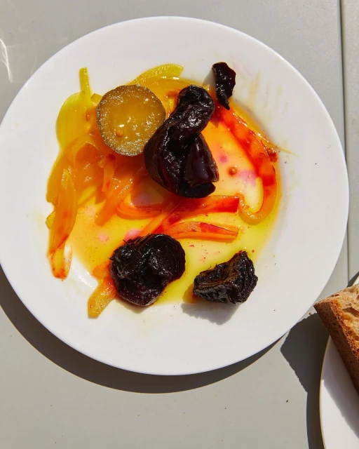 Overhead shot of a white plate with marinated plums, onions, peppers, a pickle, and sauce, next to a piece of bread on a neutral surface.