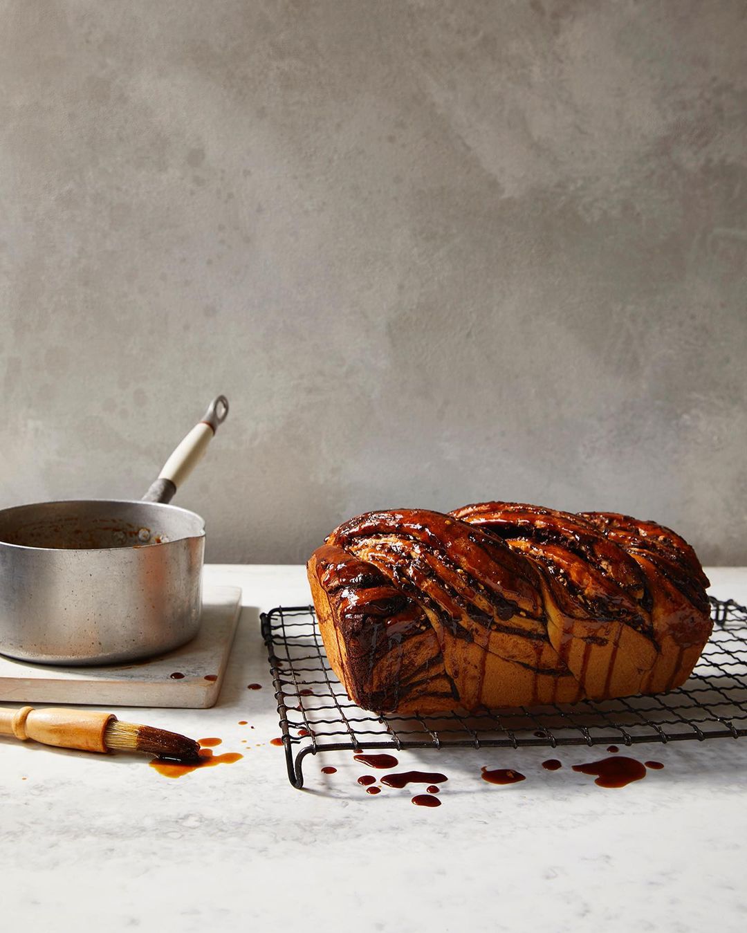 Braided loaf of chocolate babka on a wire rack, next to a pot of chocolate sauce and brush on a light coloured counter.