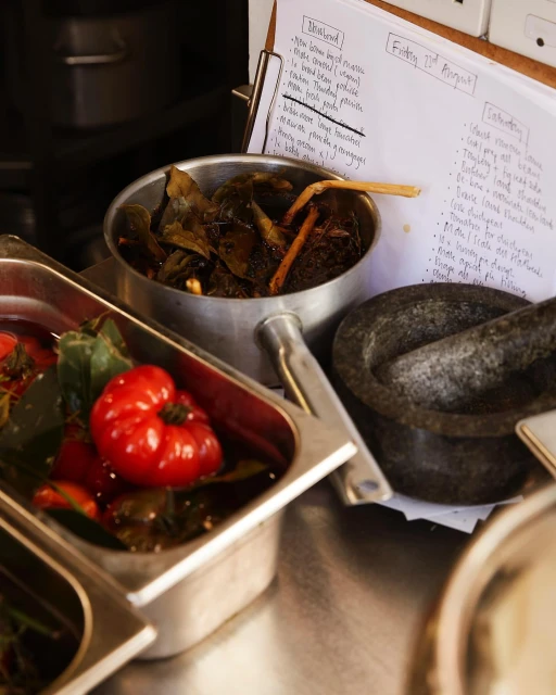 Food preparation scene: stainless steel containers with red tomatoes and green leaves, mortar & pestle, and handwritten recipe notes.