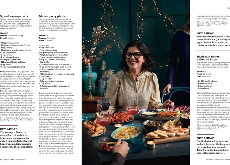Smiling woman in glasses seated at table with food, drinks, and festive lights in a warm, inviting indoor setting; holiday party.