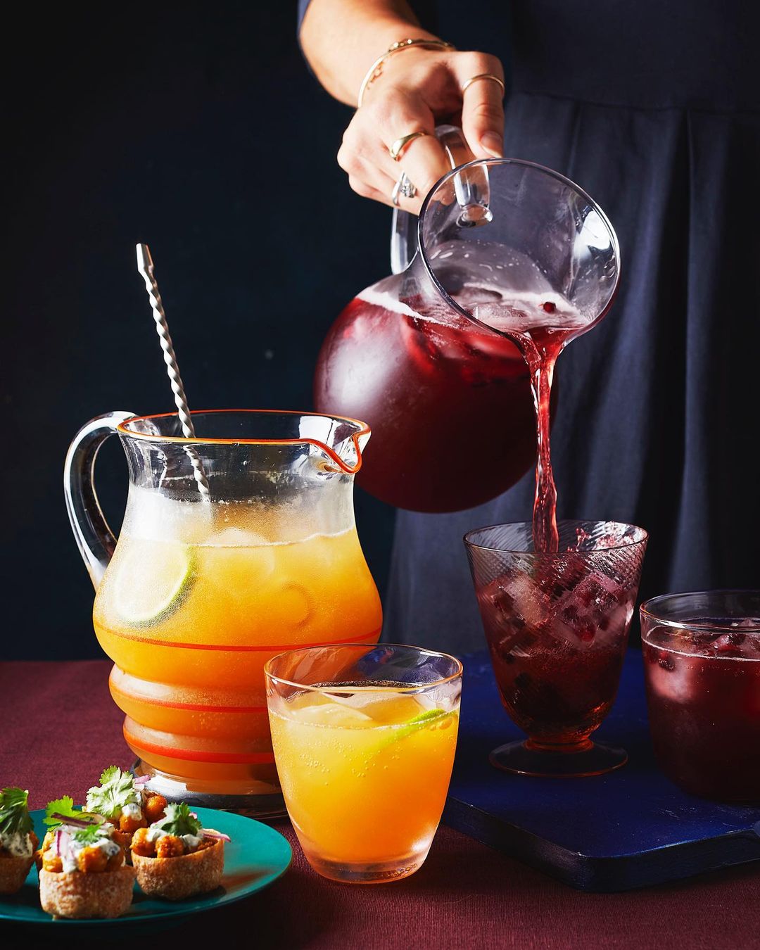 A woman is pouring dark red liquid from a clear pitcher into a glass with ice. There are several glasses and appetizers on the table.