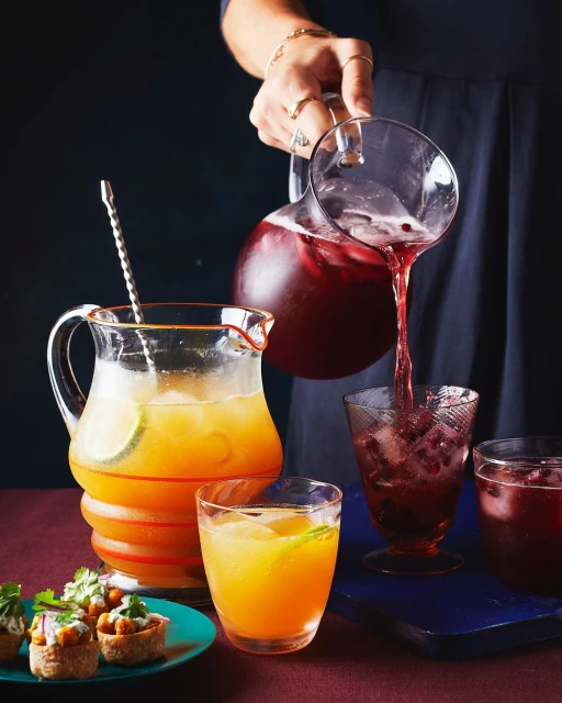 A woman is pouring dark red liquid from a clear pitcher into a glass with ice. There are several glasses and appetizers on the table.