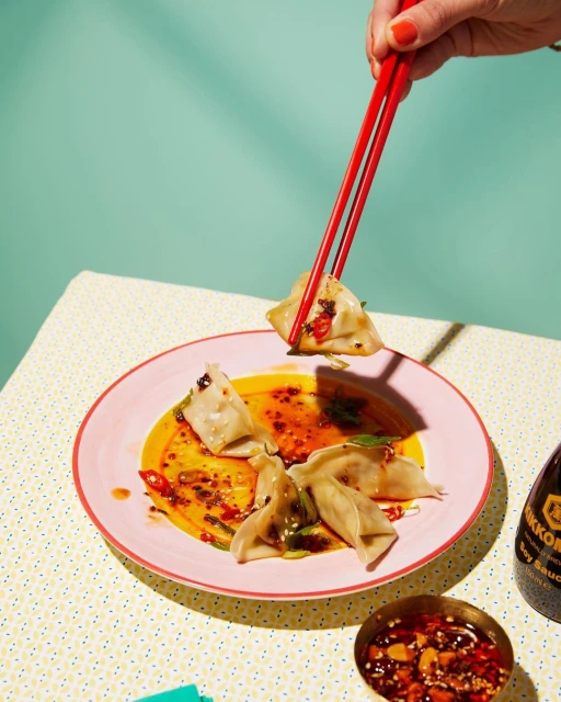 Overhead shot of dumplings on a pink plate with red chopsticks, soy sauce, and chili oil on a patterned tablecloth against an aqua wall.