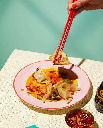 Overhead shot of dumplings on a pink plate with red chopsticks, soy sauce, and chili oil on a patterned tablecloth against an aqua wall.
