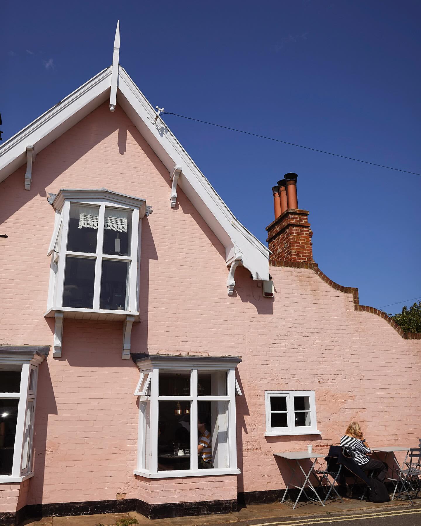 Charming pink building with white trim and windows, complemented by a blue sky. An individual sits at a table outside the building.