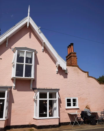 Charming pink building with white trim and windows, complemented by a blue sky. An individual sits at a table outside the building.