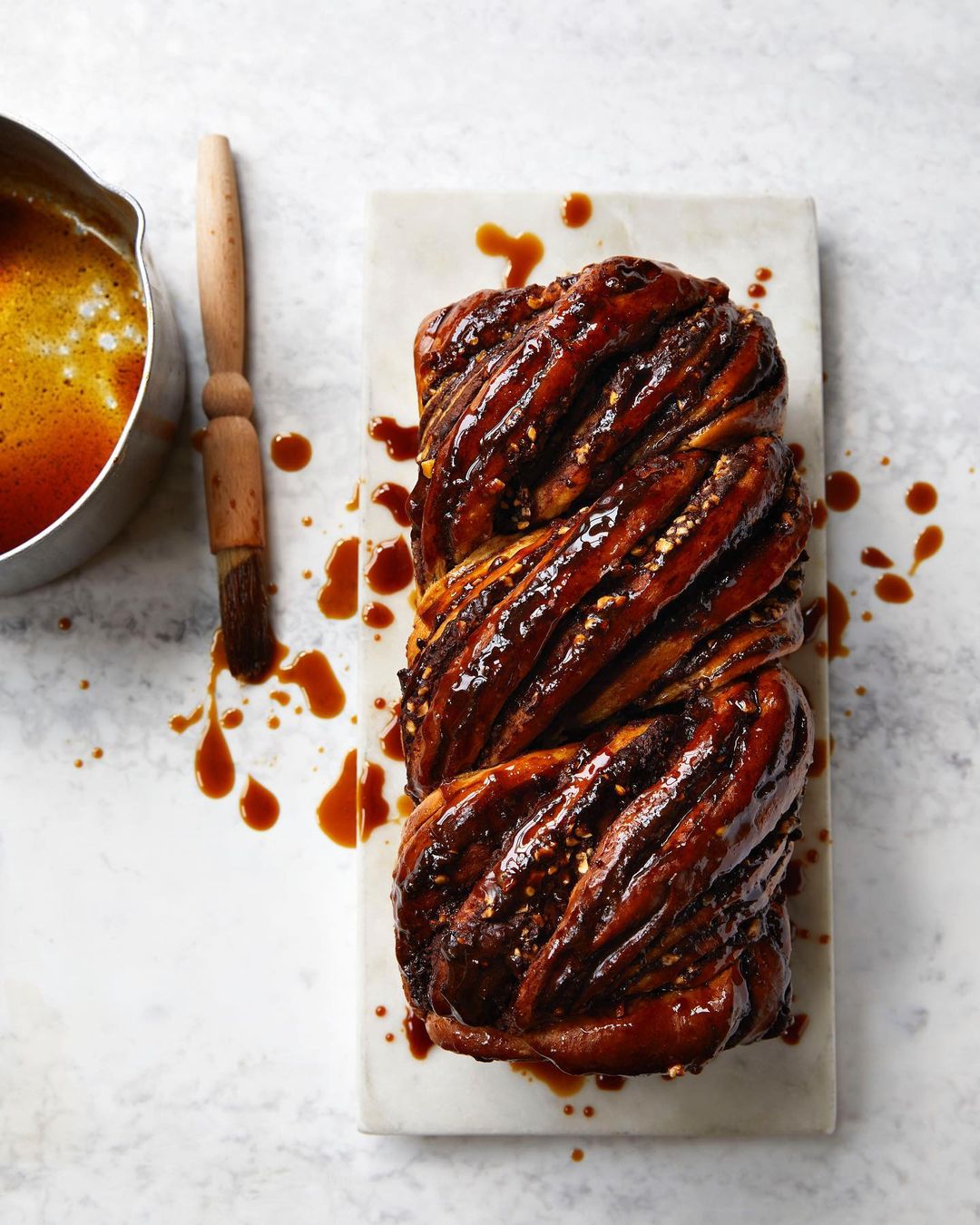 Overhead studio shot of a caramel babka bread loaf on a marble slab with a sauce brush and a bowl of warm caramel sauce.
