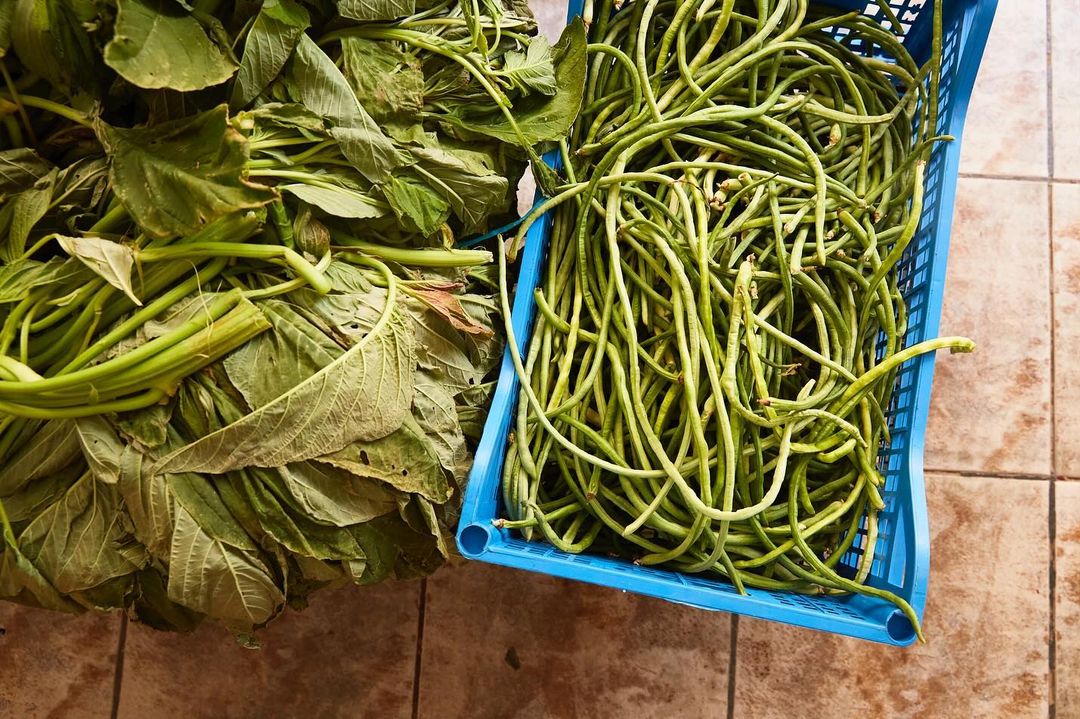 Overhead shot of leafy greens and long beans spilling from a blue basket, displayed on a tiled surface in an indoor setting.