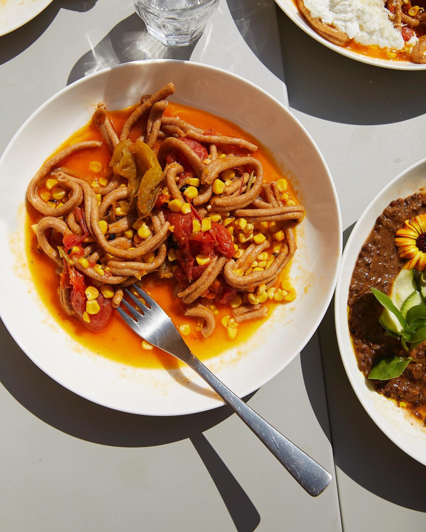 Overhead view of pasta dish with tomato sauce, corn, and peppers on a white plate next to other dishes, under bright light.