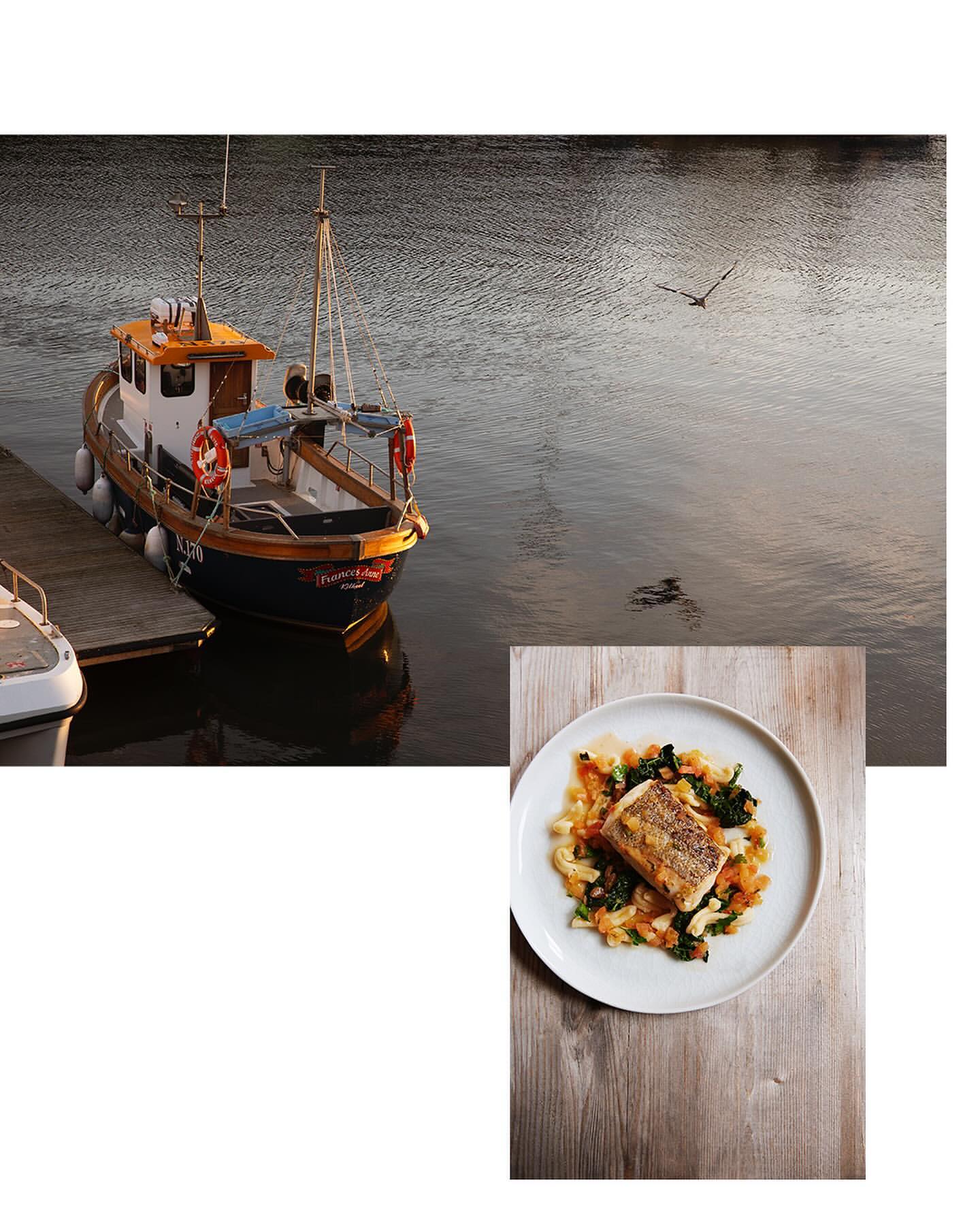 A food still life features a plate with a cooked piece of fish with vegetables on a wooden surface with a fishing boat in the background.