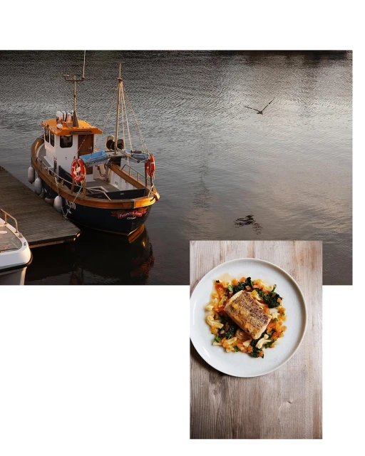 A food still life features a plate with a cooked piece of fish with vegetables on a wooden surface with a fishing boat in the background.