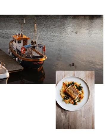 A food still life features a plate with a cooked piece of fish with vegetables on a wooden surface with a fishing boat in the background.