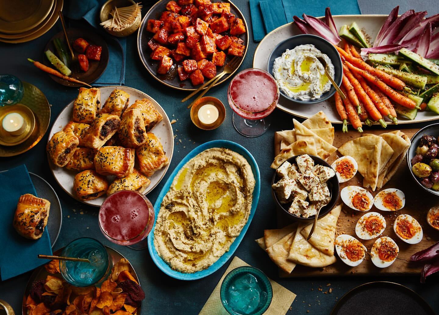 Overhead shot of a table laden with appetizers including hummus, cheese, pastries, carrots, dips, eggs, and drinks, creating a festive feast.