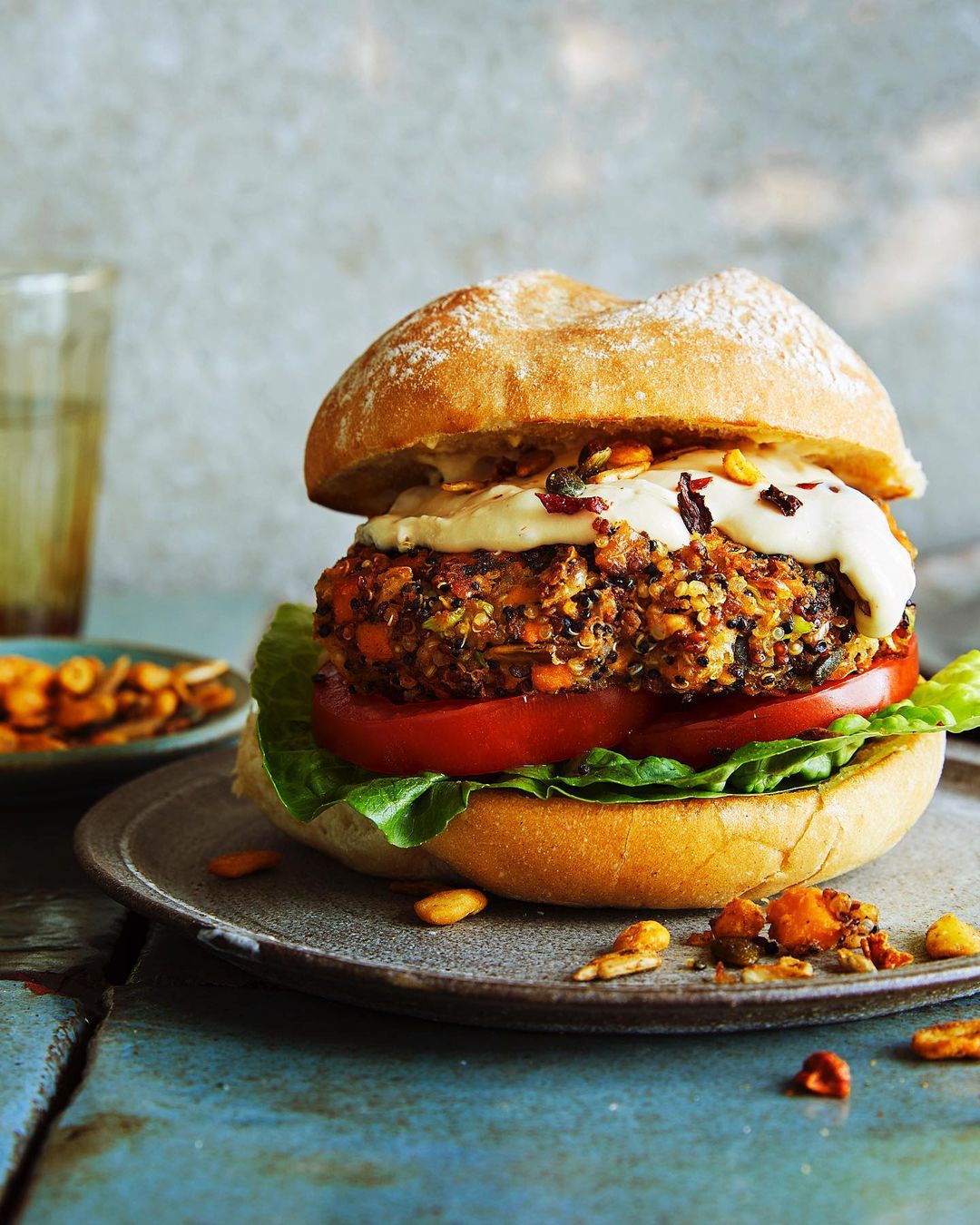 A close-up food photo showcases a veggie burger with fresh toppings and creamy sauce, set on a gray plate with scattered grains.