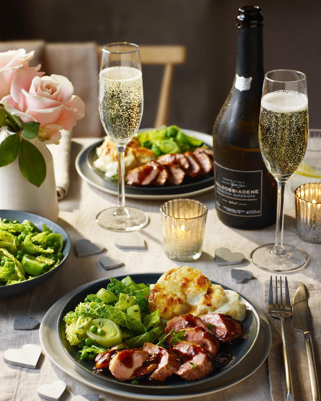 Elegant table setting featuring champagne glasses, wine, meat dishes with vegetables, candles, and pink roses, creating a celebratory ambiance.
