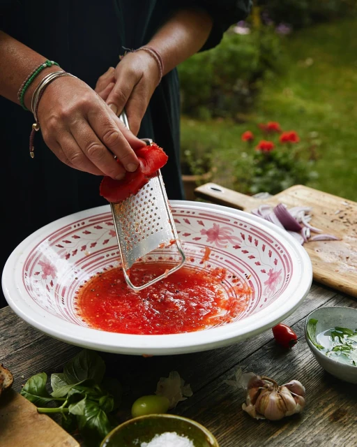 Adult is grating fresh tomatoes into a bowl on a wooden table outside. Green background with cooking ingredients visible.