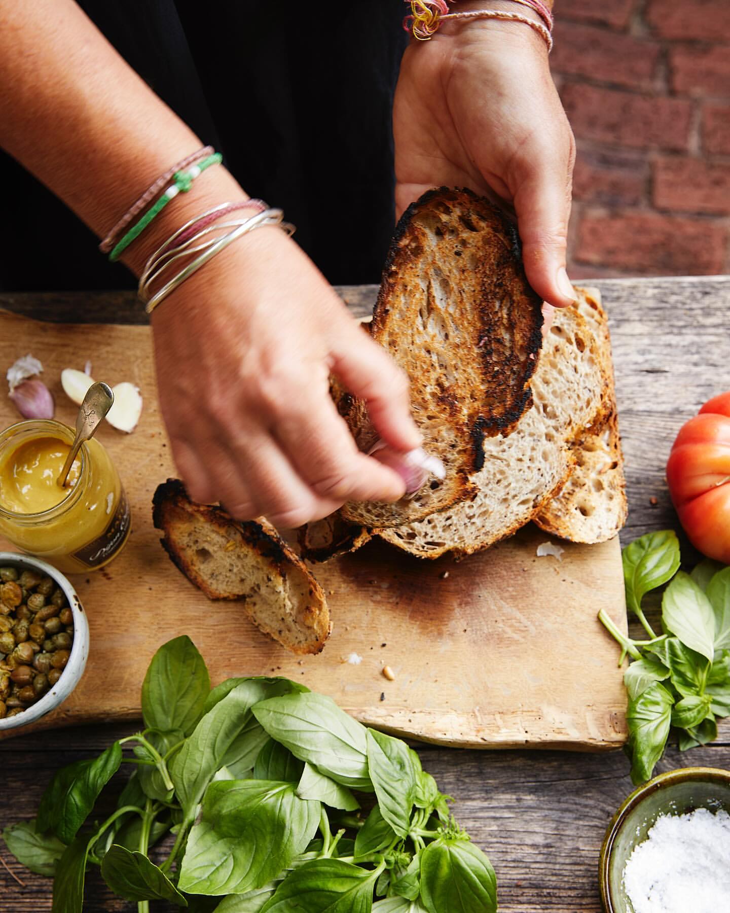 Toasted bread slices being prepared by hands on a wooden board with basil, garlic, capers, mustard, tomato, and salt.