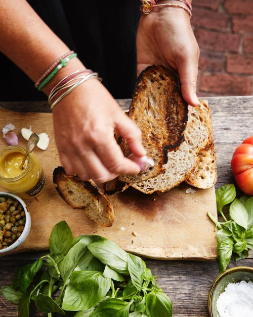 Toasted bread slices being prepared by hands on a wooden board with basil, garlic, capers, mustard, tomato, and salt.