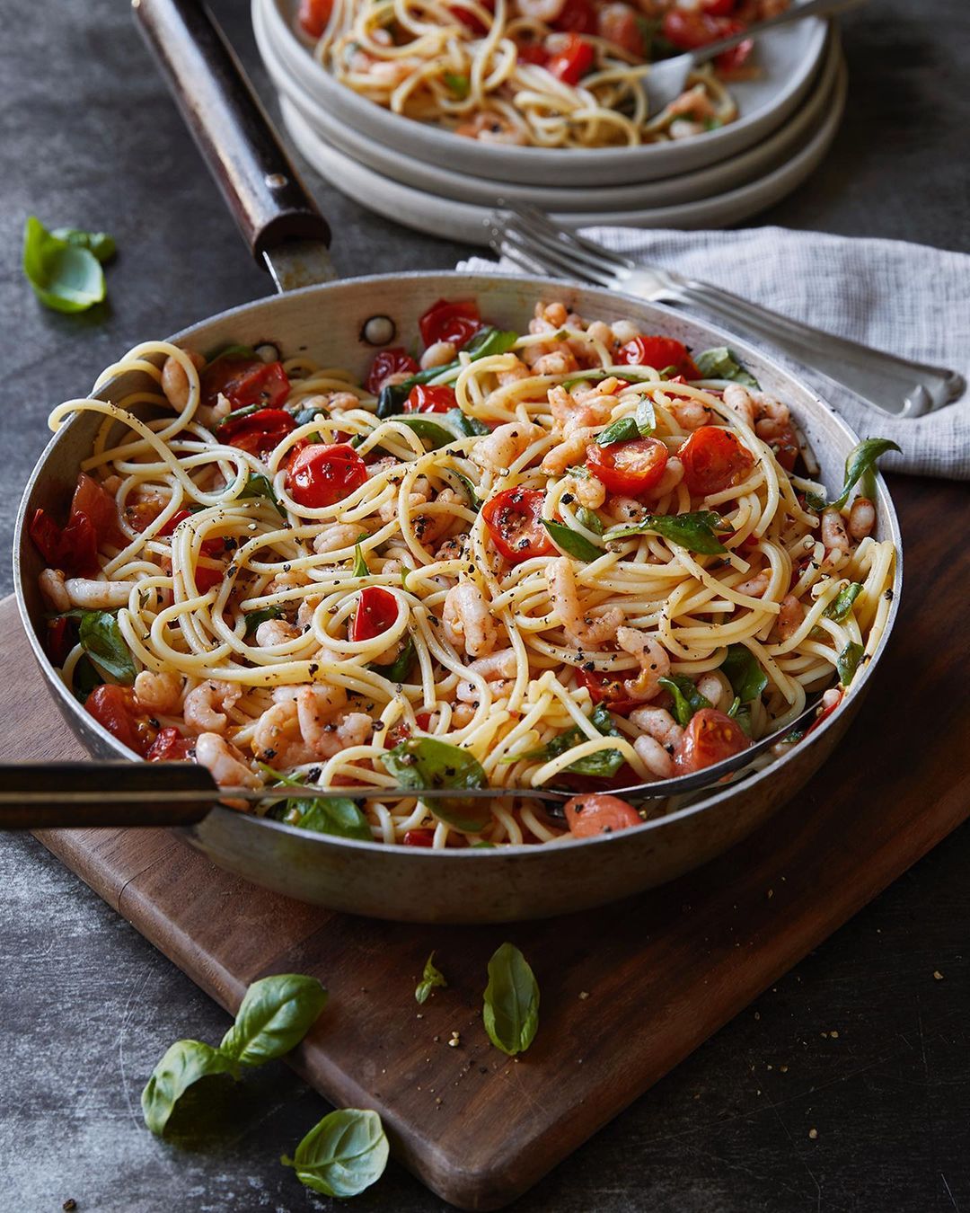 Overhead shot of spaghetti with tomatoes, shrimp, white beans, and basil in a silver pan on a wooden board, with plates and forks.