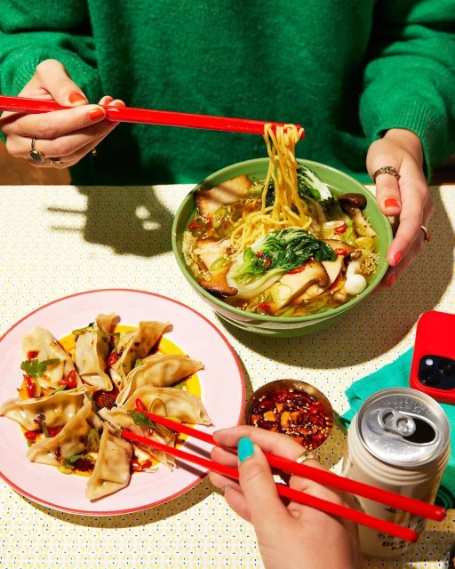 Overhead shot of two people enjoying a vibrant table setting with ramen, dumplings, chili oil, and a beverage can, perfect for a foodie feast.