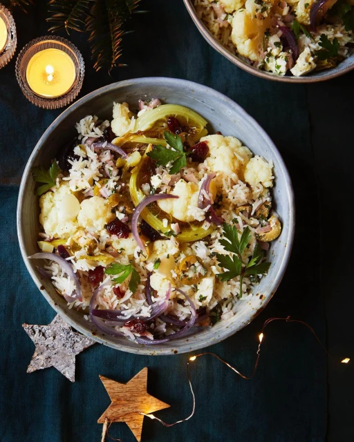 Overhead shot of a ceramic bowl filled with a cauliflower salad with rice, onions, parsley, and cranberries, sitting on a dark tablecloth.