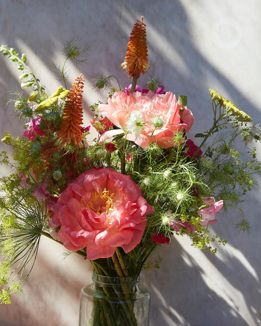 Close-up of a vibrant floral arrangement featuring pink peonies, orange kniphofia, and delicate green foliage in a clear glass vase.