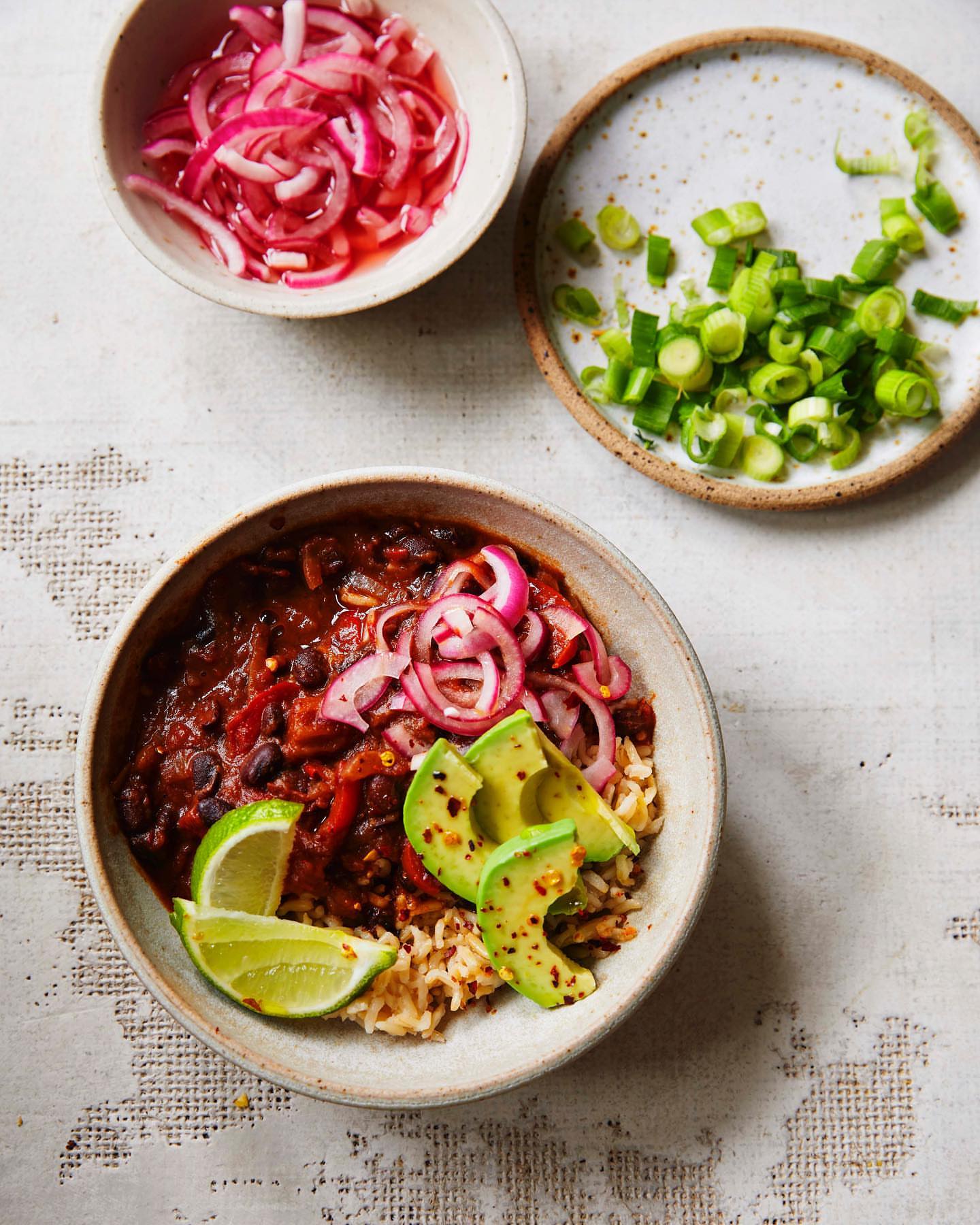 Overhead view of chili with avocado, red onion, and lime served in bowl next to small bowls of red onion and green onion.