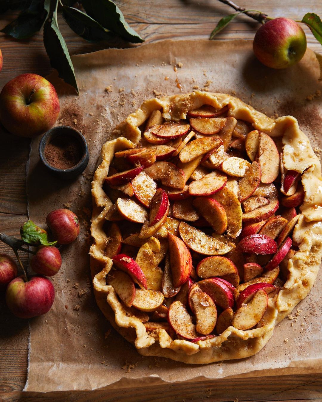 Overhead shot of an apple galette on parchment paper with a wooden table backdrop, accompanied by loose apples and leaves.