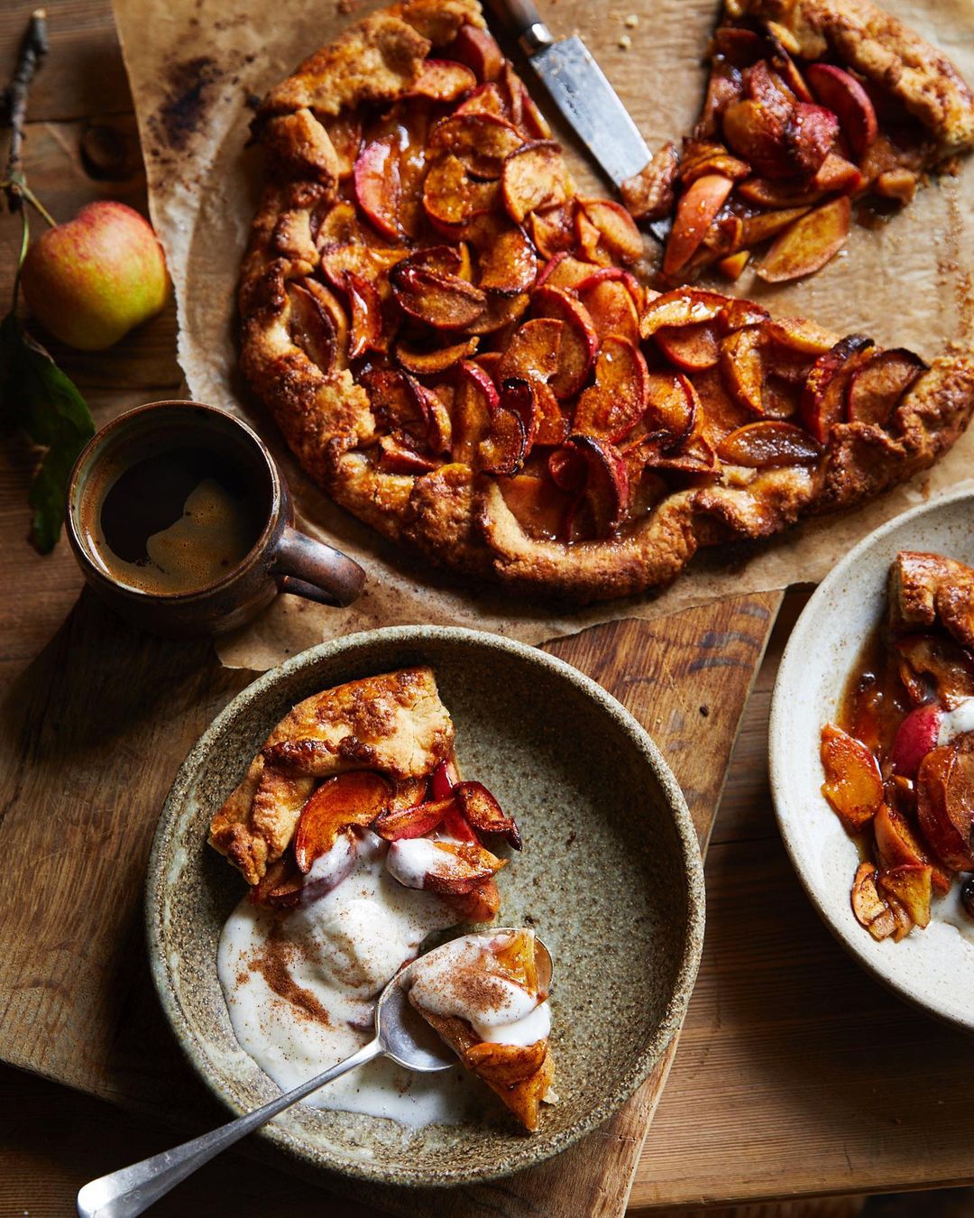 Rustic apple galette served with a scoop of ice cream on a wooden table next to a cup of coffee and an apple, shot in natural light.