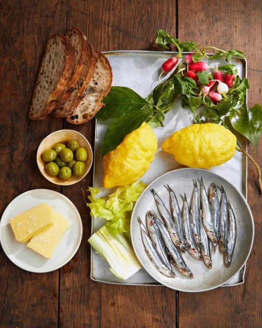 Overhead shot of a wooden table displaying a spread of sardines, lemons, olives, bread, cheese and radishes on a metal serving tray.