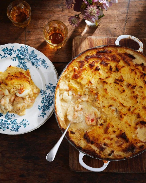 Overhead shot of fish pie in a baking dish with a slice plated on a floral dish, accompanied by liquor glasses and flowers on wood.