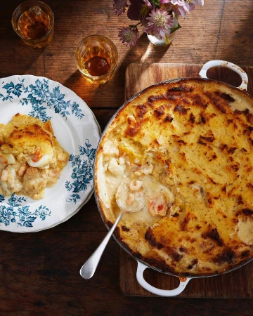 Overhead shot of fish pie in a baking dish with a slice plated on a floral dish, accompanied by liquor glasses and flowers on wood.
