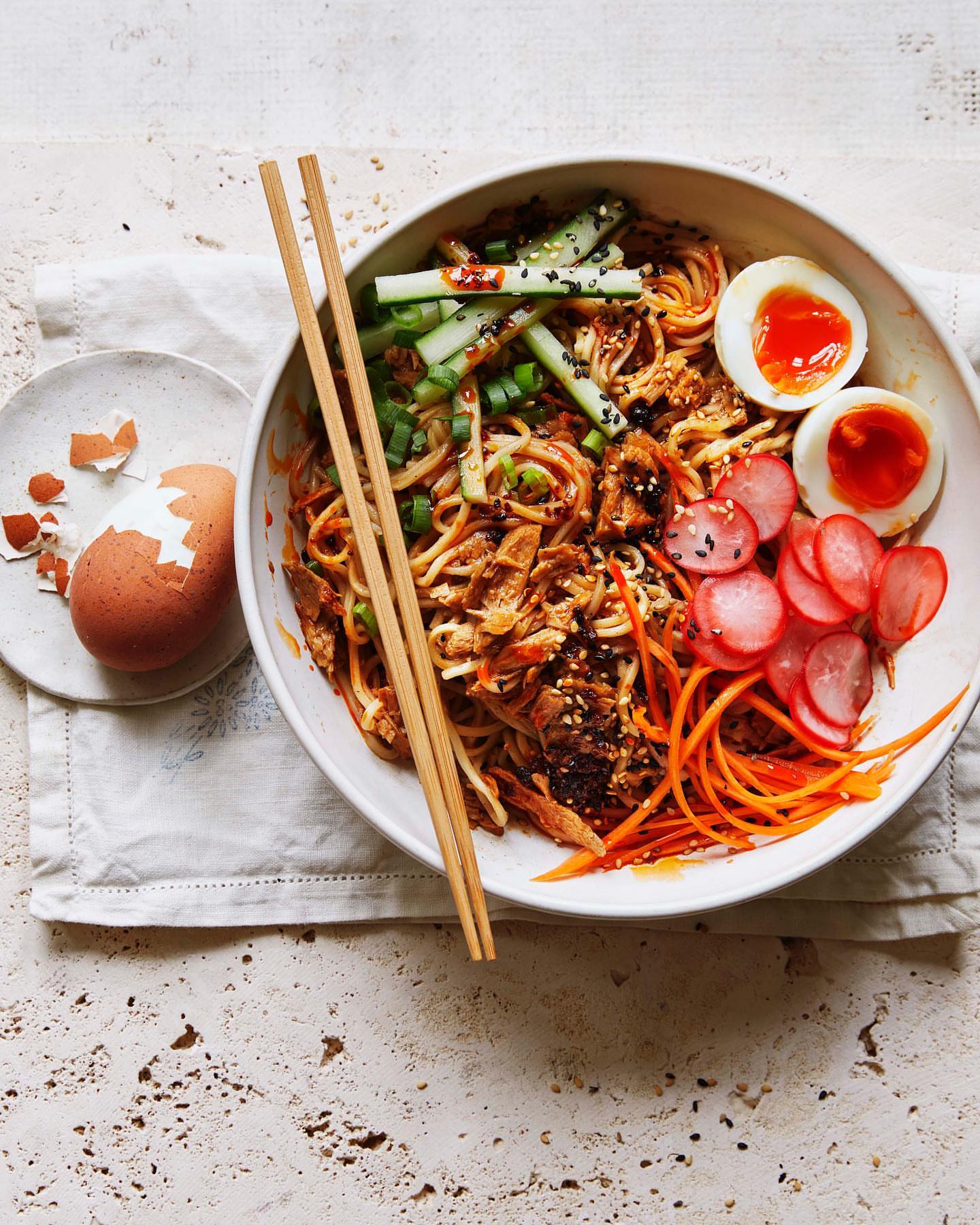 Overhead shot of a bowl of Asian noodles with chopsticks, sliced cucumbers, carrots, radishes, and a halved soft-boiled egg.