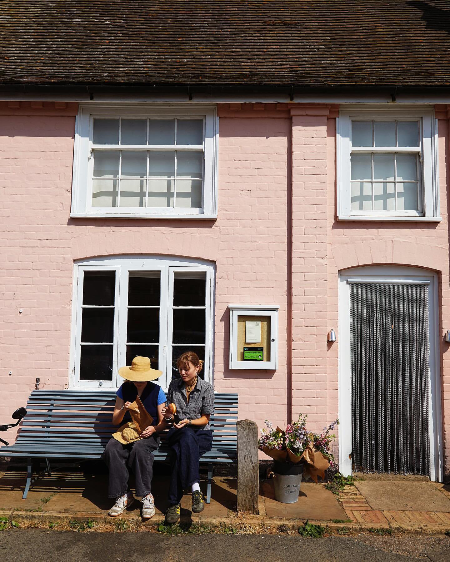 Two women relax on a bench outside a pink brick building with white-framed windows and a chainmail curtain door in the sunlight.