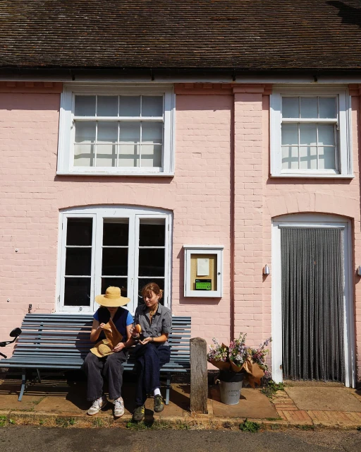 Two women relax on a bench outside a pink brick building with white-framed windows and a chainmail curtain door in the sunlight.