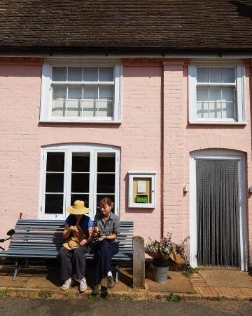 Two women relax on a bench outside a pink brick building with white-framed windows and a chainmail curtain door in the sunlight.