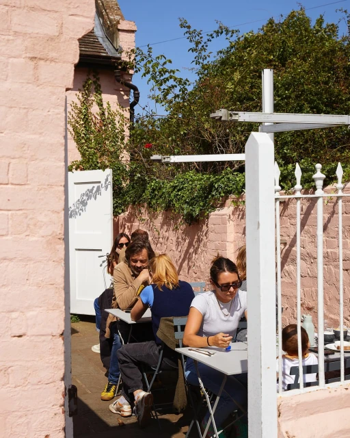 Outdoor cafe scene: Several people sit at metal tables, enjoying a sunny day amidst the pink building and lush green foliage backdrop.