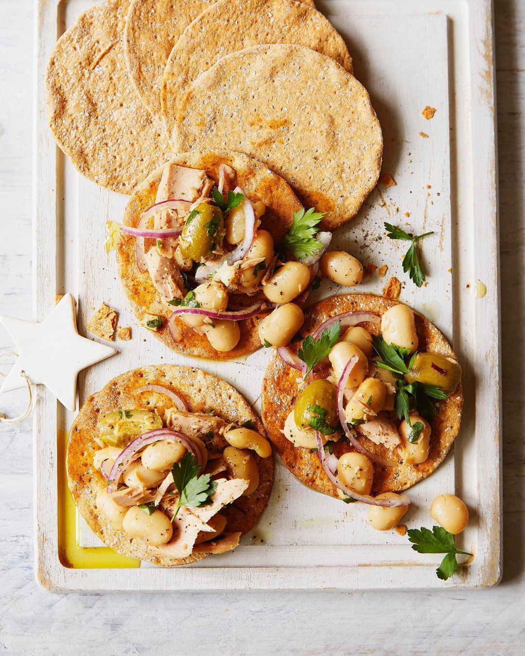 Overhead shot of three tuna and white bean tostadas on a white cutting board, garnished with red onion, parsley, and green olives.
