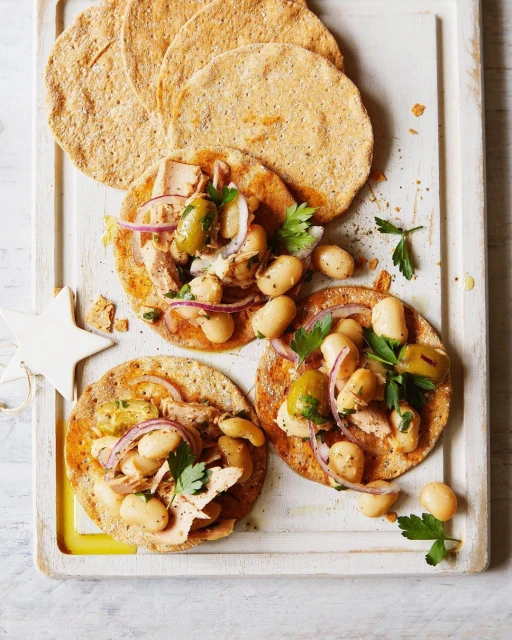 Overhead shot of three tuna and white bean tostadas on a white cutting board, garnished with red onion, parsley, and green olives.