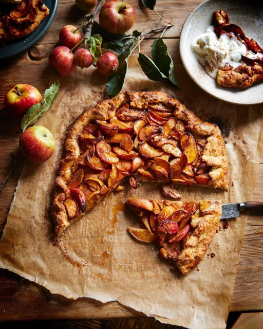 Rustic apple galette with sliced apples on parchment paper and a wooden surface, accompanied by ice cream and fresh apples.