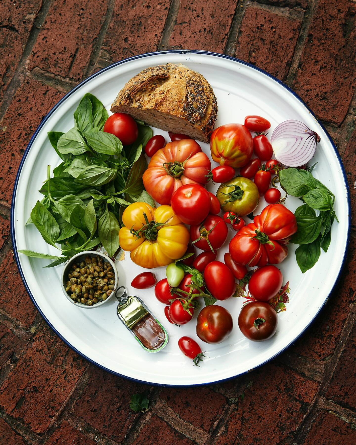 Overhead view of a white platter with heirloom tomatoes, basil, red onion, capers, loaf of bread, and a knife, on a brick surface.