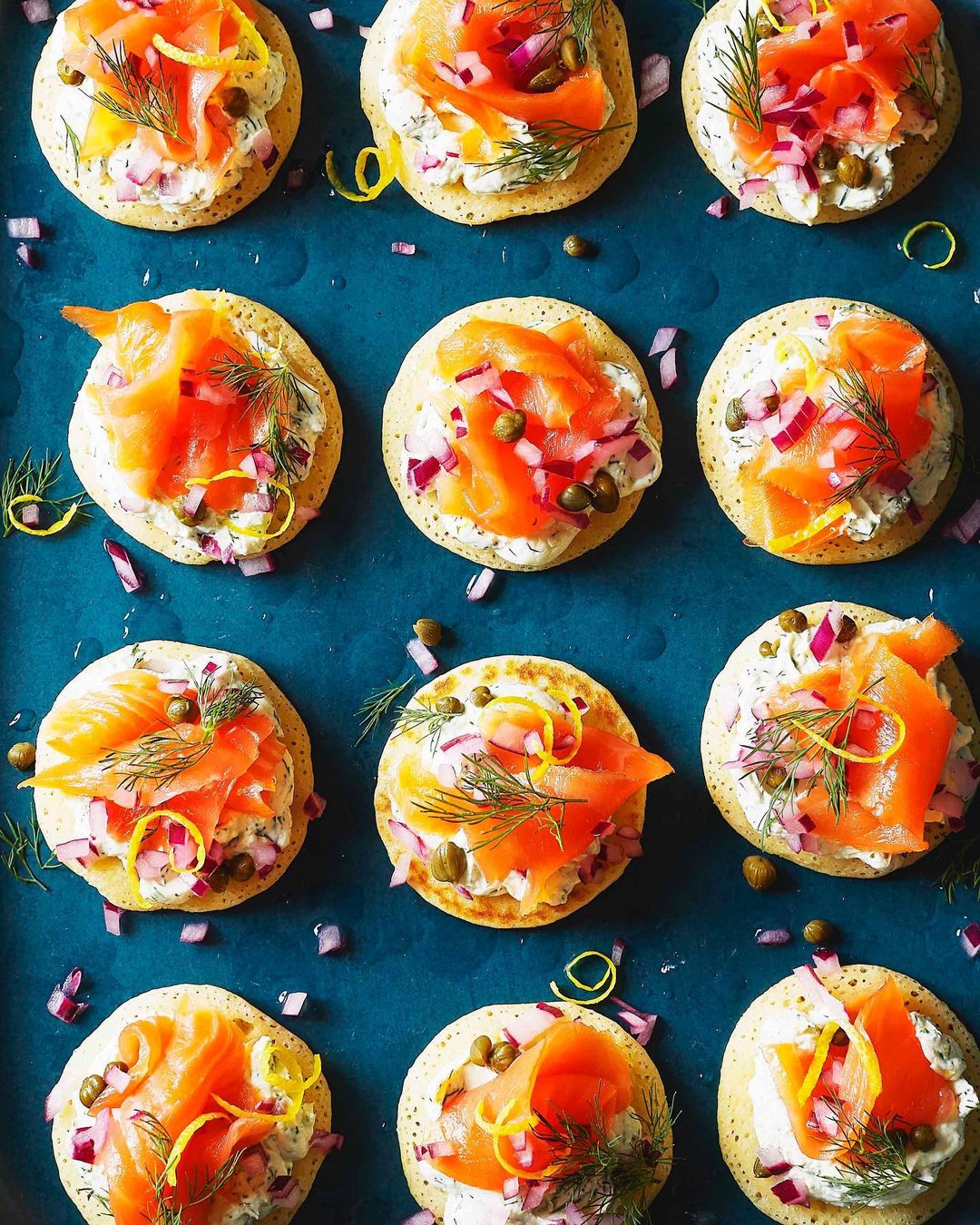 Overhead shot of canape blinis with smoked salmon, cream cheese, red onion, dill, capers, and lemon on a dark blue tray.