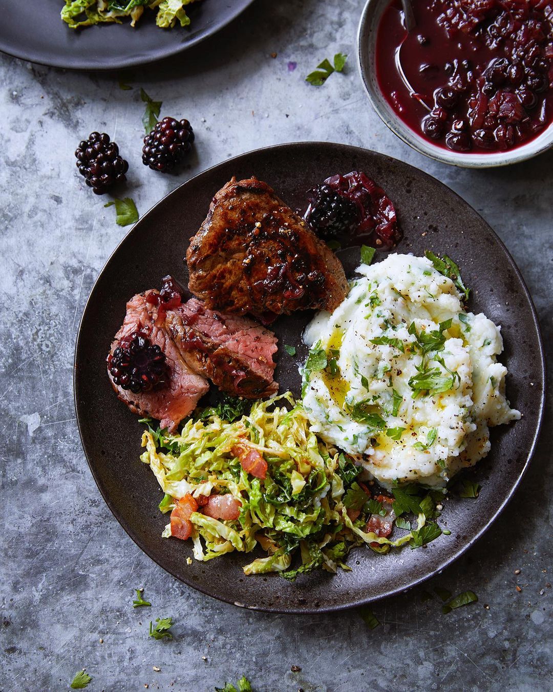 Overhead shot showcases a dark plate with steak, mashed potatoes, salad, and blackberries, set against a textured gray surface.