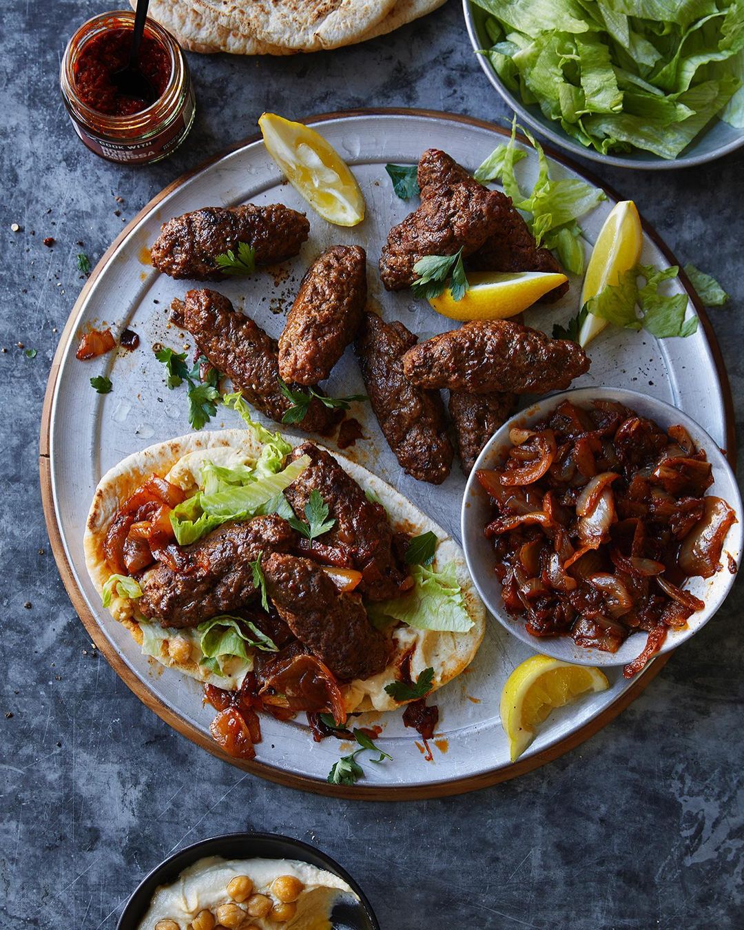 Overhead shot of kofta kebabs served on pita bread with lemon wedges, lettuce, and caramelized onions on a rustic grey background.